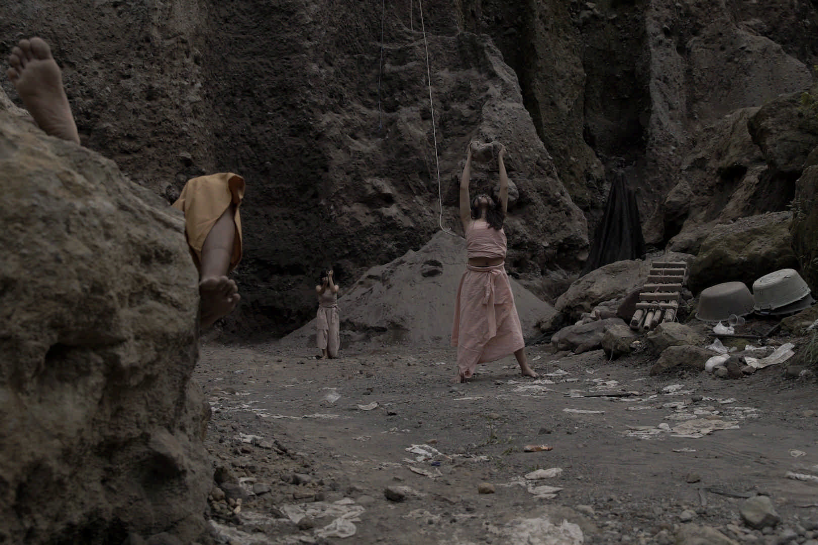 At the bottom of a dusty, earthy ravine, a person stands just off centre, they are wearing a pale pink wrap top and wrap skirt, they raise a large rock above their heads and look towards the rock. A second person dressed in a beige top and trousers raises both arms so their face is covered. A third person’s feet are visible. The soles of their feet face the camera and they appear to be lying on top of a tall rock formation. White plastic bags are trampled and strewn in the earth and a washing tub and wooden ladder-like structure are in frame.